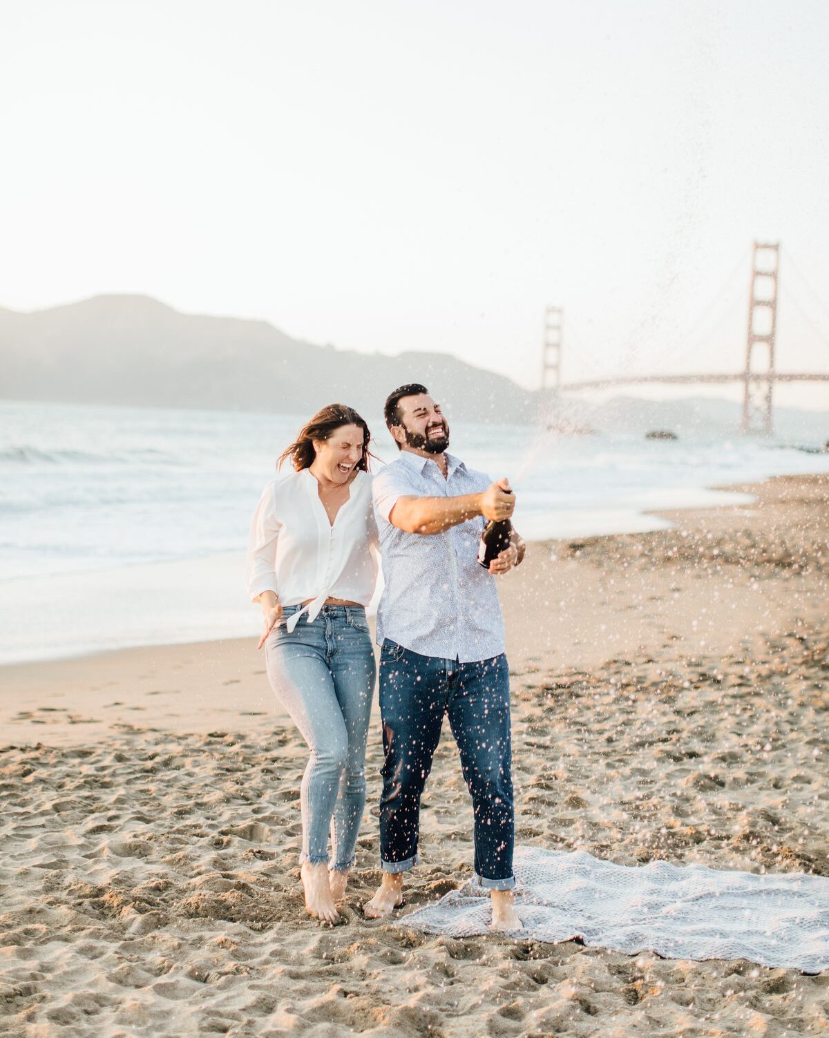 couple popping champagne together in excitement on the beach with the golden gate bridge behind them